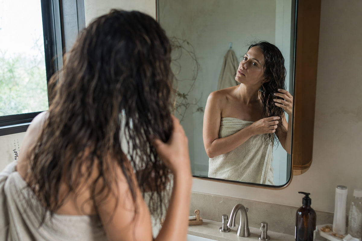Woman in a towel looking at herself in the mirror in a bathroom.
